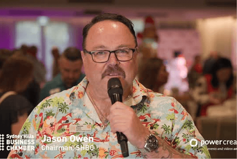 Man speaking into a microphone at an event with 'Sydney Hills Business Chamber' branding.